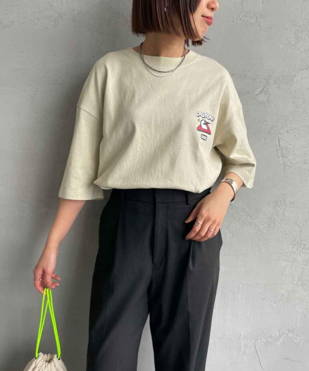 Woman wearing oversized cream graphic t-shirt with caution sign, black pants, holding bag