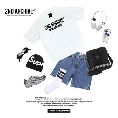 Flat lay of casual streetwear: white graphic logo tee, denim shorts, striped socks, grey sneakers, black beanie, sunglasses, black shoulder bag, and headphones.